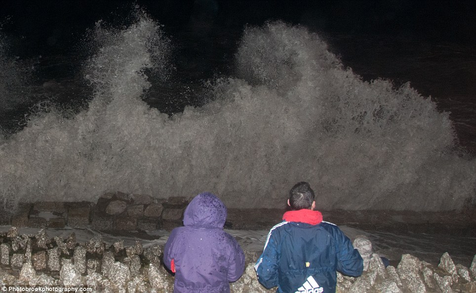 A couple stand looking at the waves crashing against a wall at Towan Beach in Cornwall at high tide
