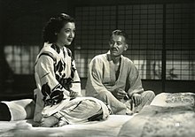 Two seated Japanese persons in traditional dress: to the left, a young woman with dark hair facing right; to the right, an elderly looking gentleman with gray hair, looking at the woman. They are sitting on futons and a shoji screen is in the background.