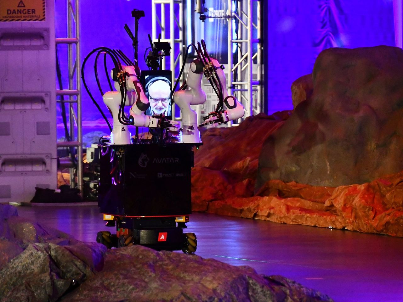 a-robot-consisting-of-two-white-arms-on-a-black-wheeled-base-drives-slowly-past-decorative-rocks-on-the-course.jpg