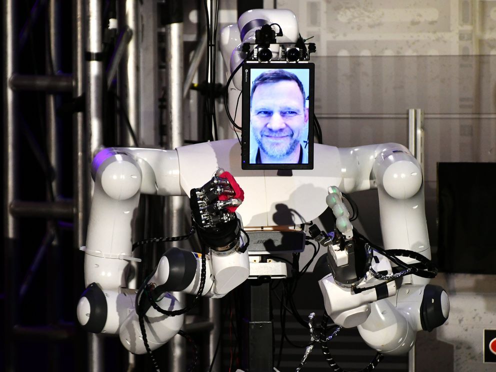 the-torso-of-a-white-robot-holds-a-red-rock-in-one-hand-with-a-picture-of-a-smiling-bearded-man-on-a-screen-where-the-robot-u20.jpg