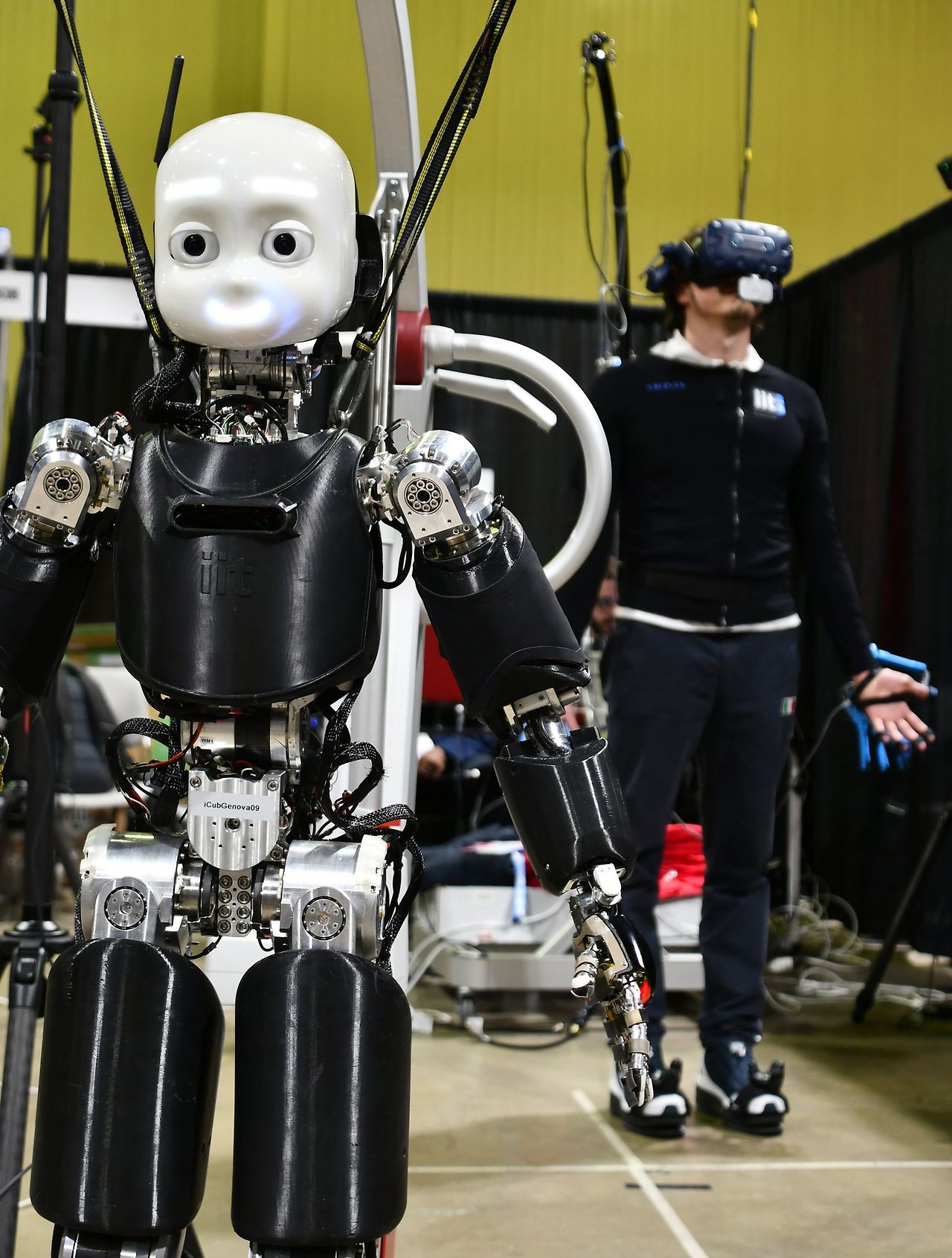 a-small-humanoid-robot-stands-in-the-foreground-while-a-human-in-a-vr-headset-stands-in-the-background-controlling-the-robot-th.jpg