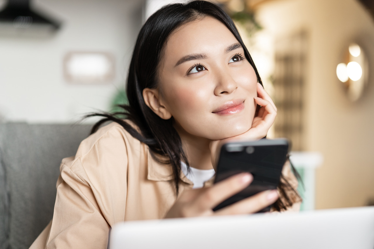 smiling-asian-girl-holding-phone-thinking-looking-up-dreamy-sitting-with-laptop-home.jpg