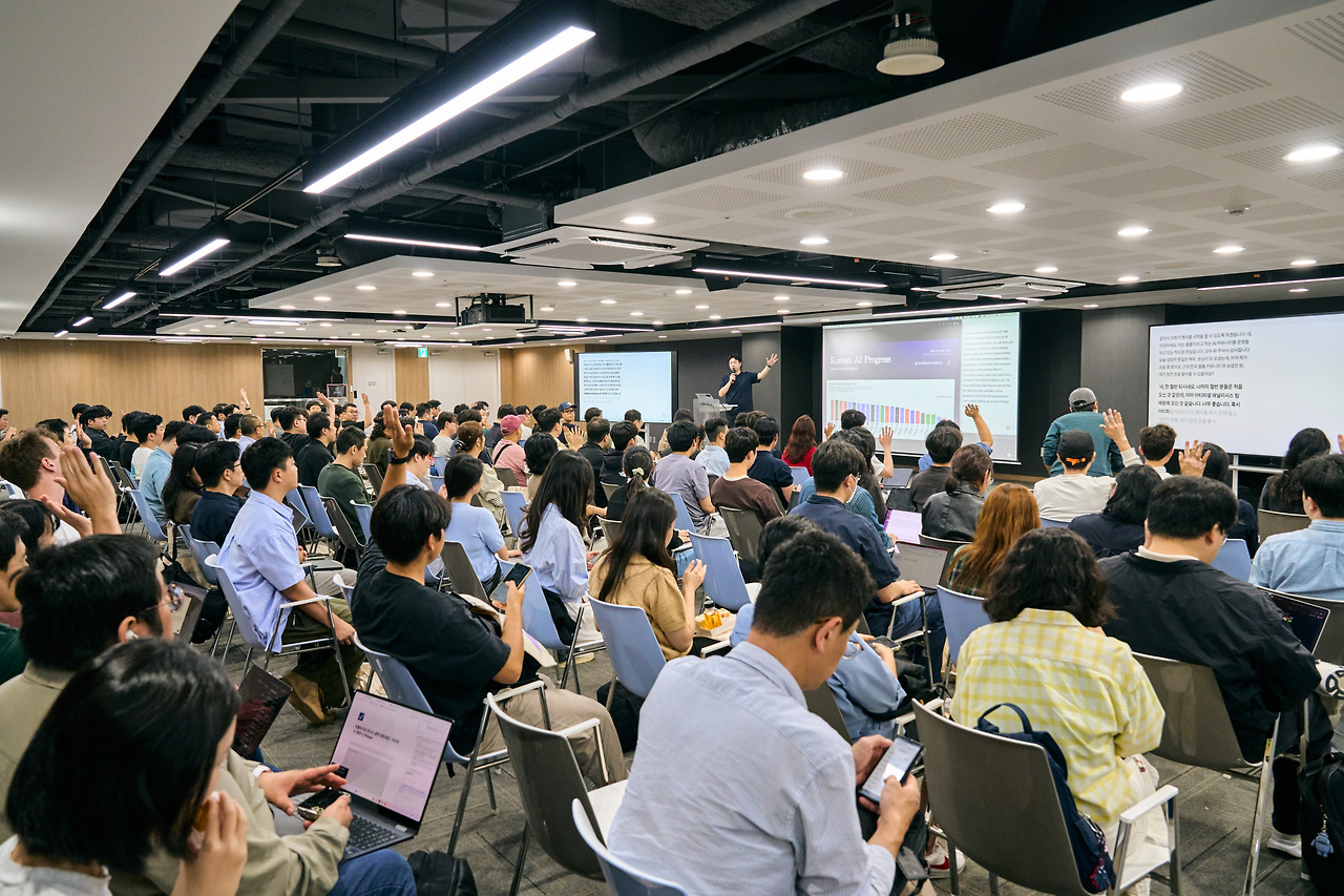 A packed lecture hall seen from the back rows, three lit screens at the front and a few raised hands.