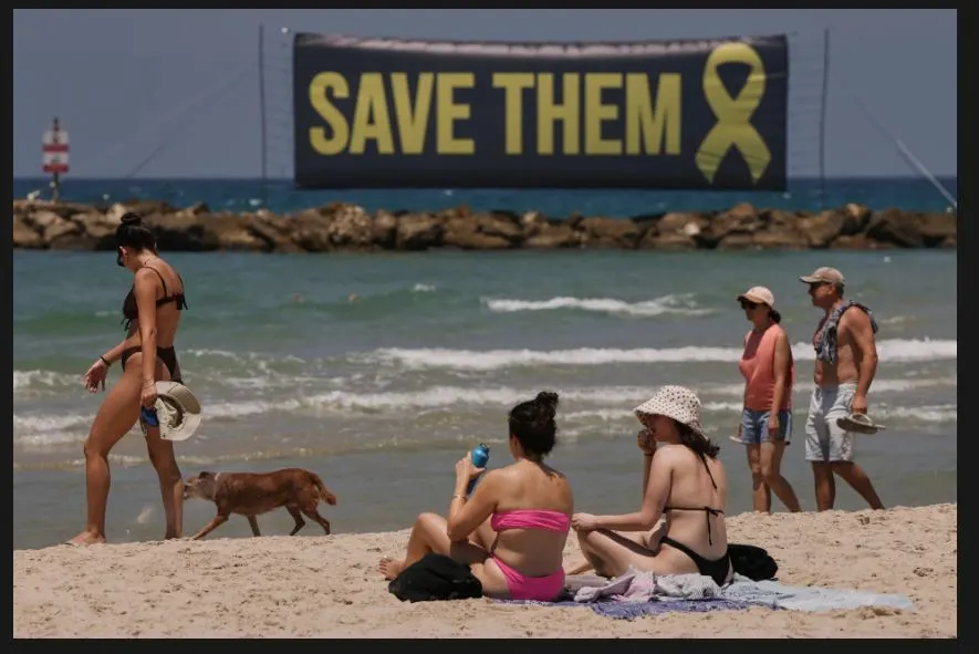 05 07 2025 Tel Aviv, Israel 04 07 2025 People sit on a beach near a banner calling for hostages held by Hamas to be saved.JPG