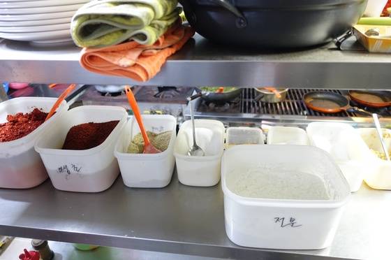 주방 한 가운데 거치대에 나란히 진열해둔 양념통 왼쪽부터 고춧가루, 매운 고춧가루, 들깻가루, 설탕, 두 가지 인공조미료, 소금, 다진 마늘, 앞에 큰 통은 전분이다. 아래층에 후추와 기름통이 보인다.
