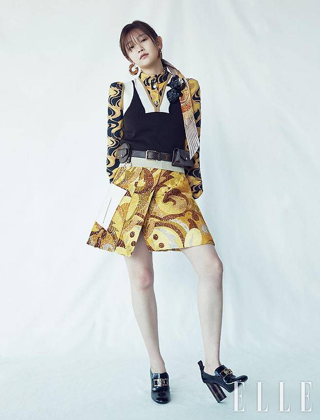 현란한 패턴의 블라우스와 A 라인 스커트, 모노톤의 베스트, 모노그램 벨트 백, 후프 이어링, 헤드 스카프, 볼드한 후프 이어링, 로고 장식의 펌프스는 모두 Louis Vuitton.