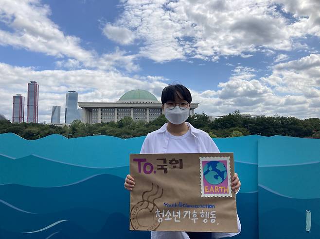 Teenage climate activist Yoon Hyeon-jeong poses for a photo in front of the National Assembly in Seoul while holding a placard urging lawmakers to take action on the climate crisis. (Yoon Hyeon-jeong)