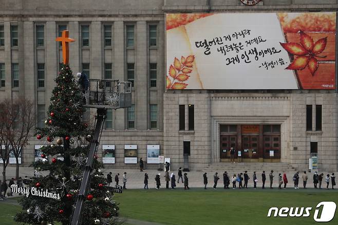 24일 오후 서울광장에 크리스마스 트리가 설치되고 있다. 설치되는 크리스마스 트리 뒤로 선별진료소 검사를 받기 위해 길게 줄을 선 시민들이 보인다. 2021.11.24/뉴스1 © News1 신웅수 기자