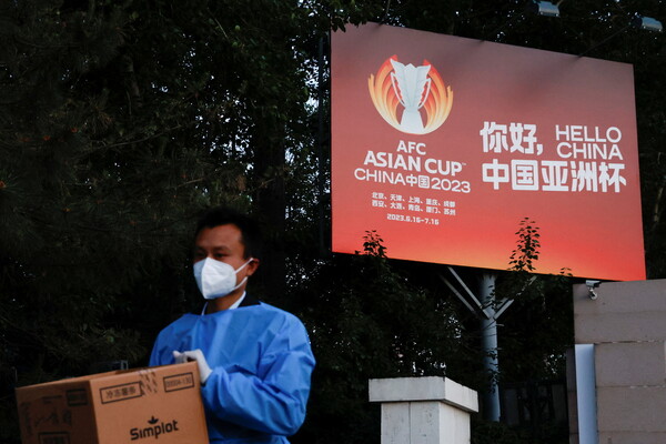 14일(현지시각) 베이징 시내에서 코로나19 방호복을 입은 사람이 2023 아시안컵을 알리는 표지판 앞을 걸어가고 있다. 베이징/로이터 연합뉴스