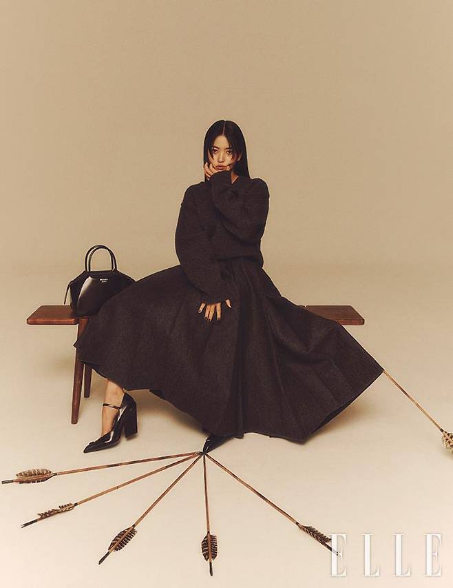 짙은 그레이 컬러 니트 톱과 풍성한 주름 장식의 풀 스커트, 로고 장식을 더한 블랙 메리 제인 슈즈, 은은한 광택의 브러시드 가죽으로 만든 토트백은 모두 Prada.