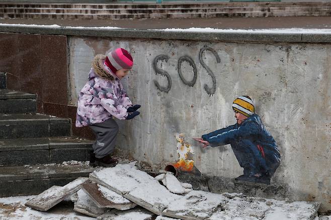 1월 29일 우크라이나 부차 거리에서 4살 어린이 아나스타샤가  이탈리아 거리 예술가 TvBoy가 그린 '불을 쬐는 소년' 그림 옆에서 불을 쬐는 시늉을 하고있다./로이터 뉴스1