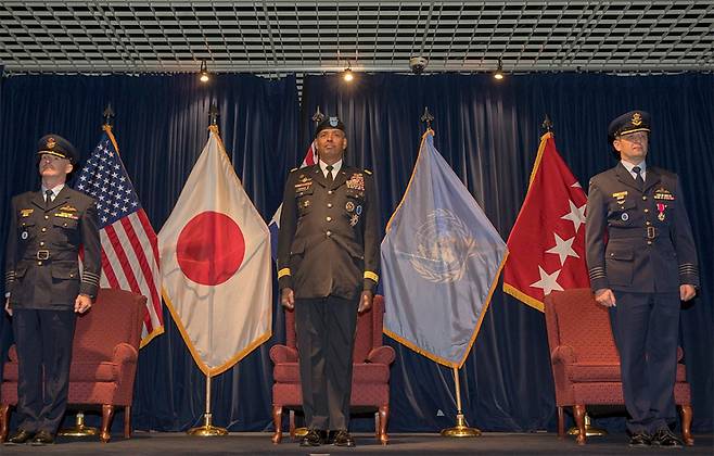 ▲ 2018년 1월 일본 요코타 공군기지에서 당시 유엔사령관 빈센트 브룩스 장군(중앙)이 일본에 있는 유엔사 후방기지 지휘관 교체 식에 참석한 모습. 3명의 군인 뒤에 놓인 미국기와 일장기, 한국 평택에 주둔한 유엔사기가 보이는데 이는 한미일 군사 연합체의 상징처럼 보인다. 사진=위키미디어