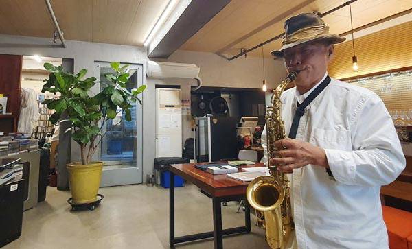 강희영 오너셰프가 그의 음식점 ‘벨 프홈나드’에서 휴식을 취하는 중 색소폰 연주를 즐기고 있다. 사진 오른쪽 중상단에 그의 애장품 하츠필드 스피커가  보인다. 강 셰프의 뒤편에는 음식점 조리대가 있다.