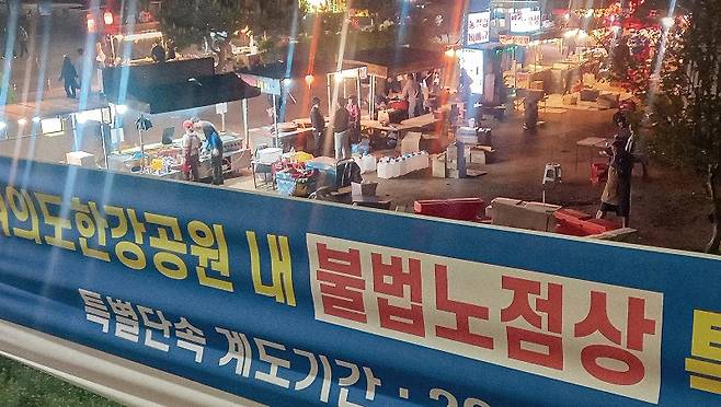 '여의도 한강공원 내 불법 노점상을 이용하지 맙시다'란 문구가 쓰여있는 현수막 옆에서 노점상이 장사를 하고 있다.[사진=더스쿠프 포토]