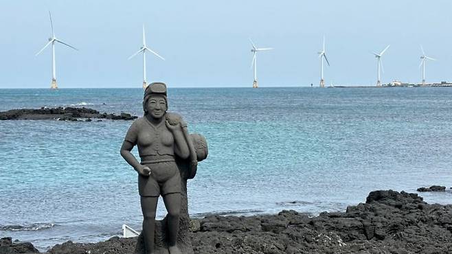 제주도 한경면 해안의 해녀 석상 뒤로 탐라해상풍력발전단지의 풍력 발전기들이 돌아가고 있다. 사진제공=연합뉴스