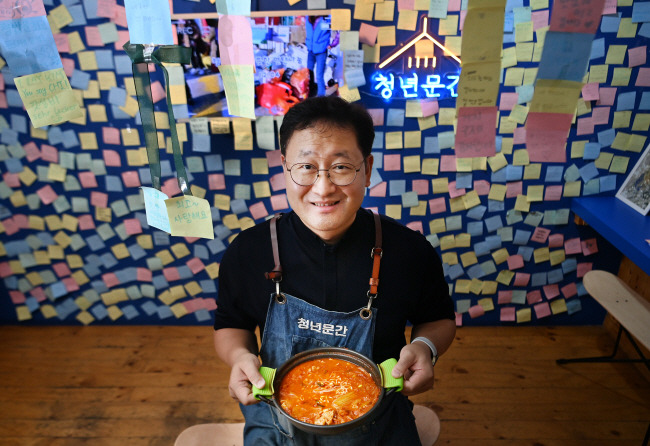 지난달 24일 ‘청년밥상문간’ 이대점에서 만난 이문수 신부. 푸짐한 1인분 김치찌개 냄비를 든 이 신부가 손님들이 붙여 둔 메모지들을 배경으로 웃고 있다. “맛있다”는 감사 인사뿐만 아니라 취업 등 개인의 소망을 쓴 경우도 있다.  윤성호 기자