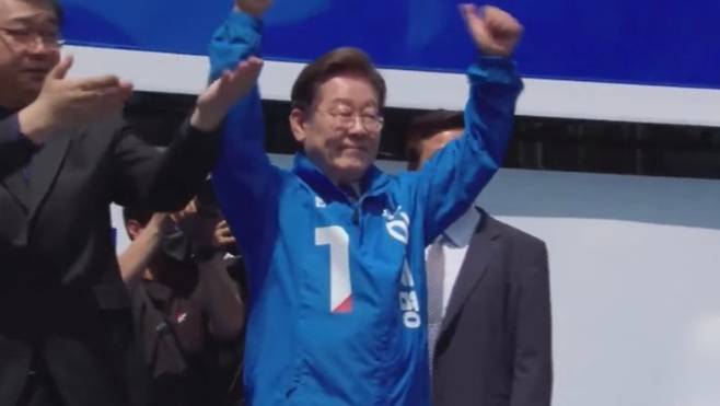 출정식 무대에 오른 이재명 더불어민주당 대선 후보가 파란 점퍼를 입고 두 팔을 들어 보이며 지지자들의 환호에 응답하고 있다.  유세 현장에는 안전 확보를 위한 방호 조치가 대폭 강화됐다. ('델리민주' 캡처)
