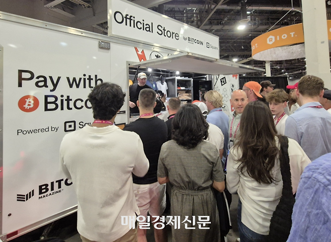 비트코인 2025 기념품샵에서 비트코인으로 기념품을 구입하기 위해 줄을 선 사람들.  비트코인으로 결제를 할 수 있다는 문구가 좌측에 크게 써 있다. [이종화 기자]