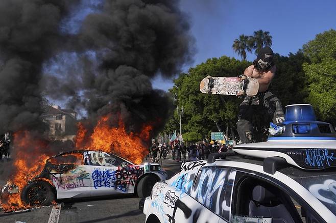 8일(현지 시간) 미 캘리포니아주 로스앤젤레스 도심 메트로폴리탄 구치소 인근에서  무인 자율주행 택시 웨이모가 불타는 가운데 한 시위 참가 남성이 또 다른 차량을 훼손하고 있다. 2025.06.09. [로스앤젤레스=AP/뉴시스]