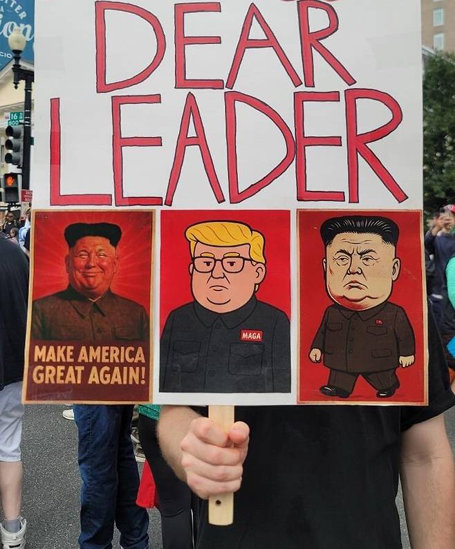 14일(현지시간) 워싱턴 DC에서 열린 미 육군 창설 250주년 기념 열병식을 앞두고 백악관 인근 라파예트 광장에서 반(反)트럼프 시위가 열렸다. 한 시위 참가자가 트럼프 대통령과 김정은 북한 국무위원장의 얼굴을 \'\'합성\'\'한 만화가 그려진 피켓을 들고 있다. 연합뉴스