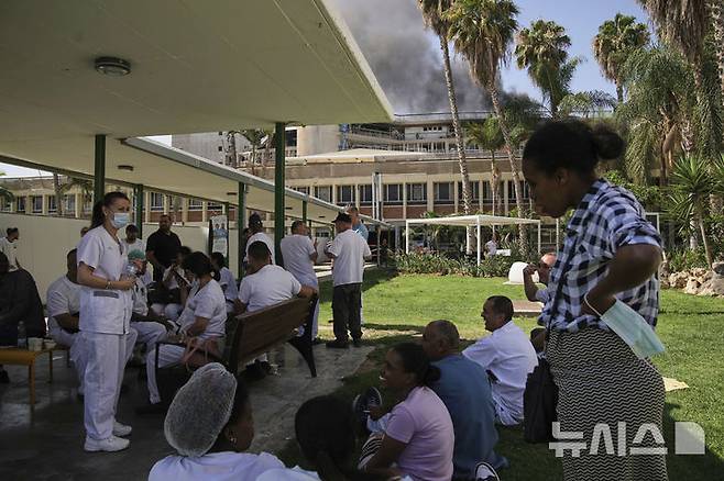 [베르셰바=AP/뉴시스] 19일(현지 시간) 이스라엘 베르셰바에 있는 소로카 병원 단지 내 건물이 이란의 미사일 공격을 받아 연기가 피어오르는 가운데 병원 밖에 의료진이 모여 있다. 이란 혁명수비대는 “병원 근처 이스라엘군 지휘·정보센터가 타격받았다”라고 주장했다. 2025.06.20.