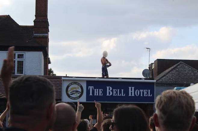 14일(현지시간) 영국 에식스주 에핑 중심가에 있는 3성급 호텔 ‘더 벨’ 앞에서 반이민 단체가 시위를 벌이고 있다. 이 호텔 위에는 한 남성이 올라가 있다. 출처=이브닝 스탠더드