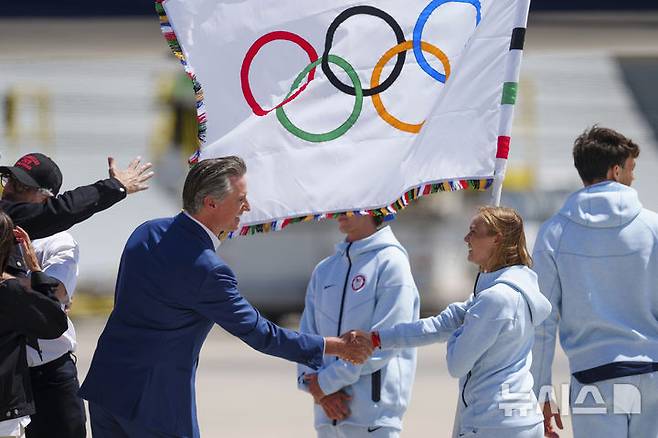 [로스앤젤레스=AP/뉴시스] 개빈 뉴섬(왼쪽) 미국 캘리포니아 주지사가 12일(현지시각) 올림픽 대회기를 들고 LA 국제공항에 도착한 일부 올림픽 선수단을 맞이하며 악수하고 있다. 2024.08.13.