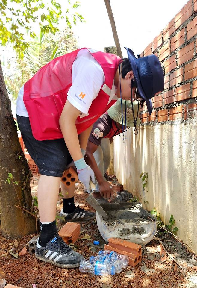 한 봉사단원이 캄폿영문 교회의 낡은 담벼락을 보수하기 위해, 현지 기술자의 도움을 받아 시멘트를 바르고 벽돌을 쌓는 작업을 하고 있다.