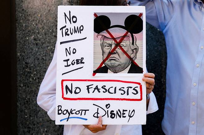 19일 뉴욕 맨해튼에 있는 에이비시 방송사 스튜디오 앞에 한 시위자가 지미 키멀 쇼 중단 결정을 비판하는 손팻말을 들고 서 있다. 도널드 트럼프 대통령의 얼굴에 미키마우스 머리띠를 그려넣은 뒤 ‘독재자 반대’ ‘디즈니 불매’ 등 문구를 써 넣었다. 에이비시 방송사는 월트디즈니컴퍼니가 소유하고 있다. 뉴욕/로이터 연합뉴스