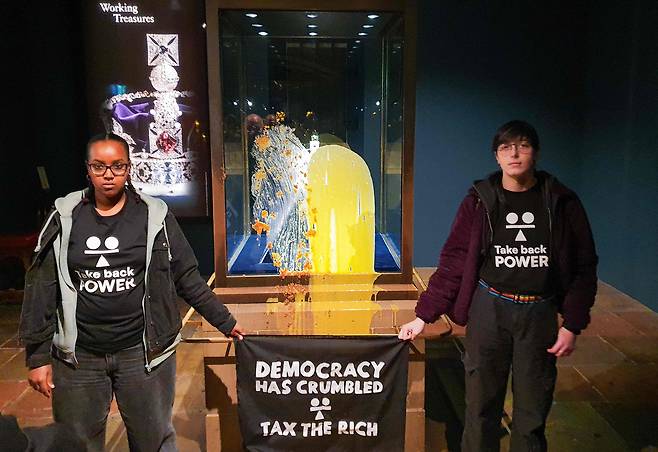 영국의 시민단체 '권력을 되찾자(Take Back Power) 회원들이 6일 영국 런던탑의 왕실 보석 전시실에서 '제국 왕관' 진열장에 음식물을 투척한 뒤 "민주주의는 무너졌다. 부자들에게 세금을 걷어라"라고 적힌 깃발을 들고 사진을 찍고 있다. /AFP 연합뉴스