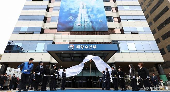 [부산=뉴시스] 최동준 기자 = 이재명 대통령이 23일 부산 동구 해양수산부 청사에서 참석자들과 함께 현판 제막식을 하고 있다.   오른쪽부터 해수부 이석민 주무관 모친 정성미 씨, 박은혜 해수부 주무관, 이희숙 원조콩나물비빔밥 사장, 이대규 SK해운 선장, 박형준 부산시장, 김성범 해수부 장관 직무대행, 이재명 대통령, 김민석 총리, 구본민 한국해양대학교 학생, 박혜라 씨드(SEA.D) 대표, 윤경희 에이치라인해운 2등 항해사, 윤병철 해수부 주무관. (대통령실통신사진기자단) 2025.12.23. photocdj@newsis.com /사진=