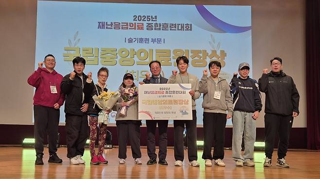 2026년 재난응급의료 종합훈련대회 단체사진으로, 여러 사람이 모여 상장과 꽃다발을 들고 엄지 척 포즈를 하고 있다.배경에는 ‘슬기로운 의술 부분! 으뜸 중앙의료 위상’이라는 문구가 적혀 있다.