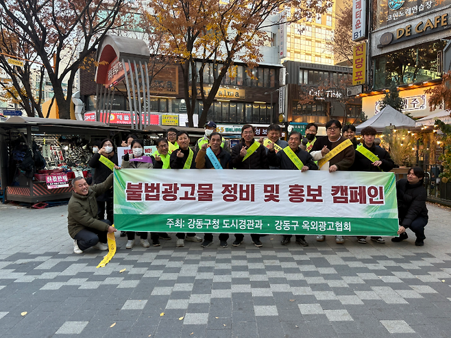 사람들이 “불법광고물 정비 및 홍보 캠페인, 주최: 강동구청 도시경관과, 강동구 옥외광고협회”라고 적힌 현수막을 들고 있는 사진이다. 캠페인 참여자들이 어깨띠를 두르고 캠페인에 참여하고 있다.
