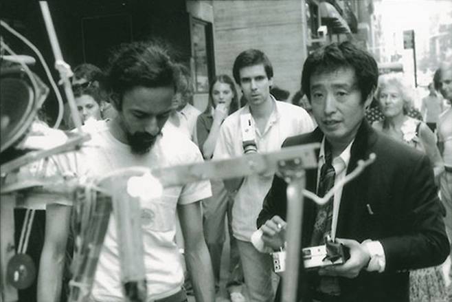 In June 1982, Nam June Paik, right, remotely operates ″Robot K-456″ (1964) on the street outside the Whitney Museum of American Art in New York. The 18-year-old robot was about to stage a mock traffic accident performance. [NAM JUNE PAIK ART CENTER ARCHIVE]