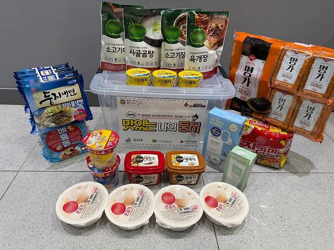 여러 종류의 음식과 생필품이 투명한 플라스틱 상자 주변에 놓여 있는 사진이다. 상자에는 ‘맛있는 나의 둥지!’라는 문구가 적혀 있다.
