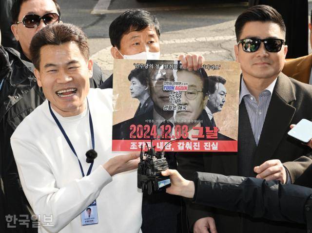 한국사 강사 출신 보수 유튜버 전한길(앞줄 왼쪽)씨가 12일 서울 동작경찰서에 피의자 신분으로 출석하면서 기자들의 질문에 자신이 제작한 영화 포스터를 보여주며 홍보를 하고 있다. 오른쪽은 김현태 전 특수전사령부 707특수임무단장(대령). 왕태석 선임기자