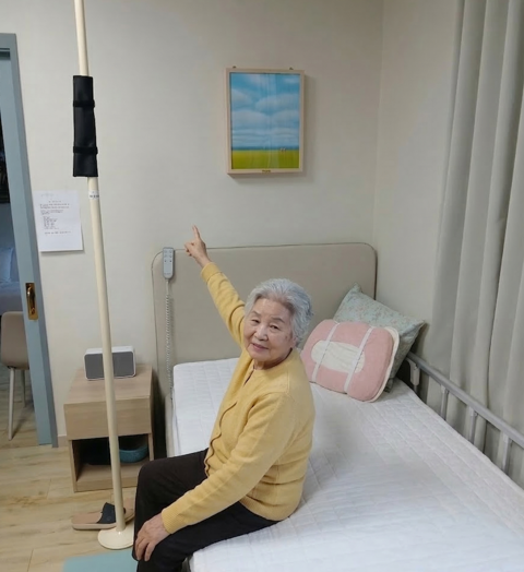 케어링스테이 입주 어르신이 객실 벽면에 설치된 홈플릭스의 '아우름 바이탈케어'를 가리키고 있다. 원목 프레임 안에 김주희 작가의 '하늘과 들판'이 담긴 이 액자형 설치물은, 웨어러블 기기 착용 없이도 심박·호흡·체동을 24시간 실시간으로 감지하는 비접촉 건강 모니터링 솔루션이다. (사진=홈플릭스 제공)