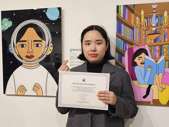 자폐 스펙트럼 장애를 지닌 제주 청년 예술가 김현정(31)이 오는 4월 10일부터 5월 22일까지 미국 뉴욕 뉴저지 민권센터(MK Space Gallery)에서 초대 개인전 《Beyond the Star: Hello, World!》를 개최한다. 앞서 지난 2일부터 8일까지는 뉴욕 맨해튼 첼시 지역의 코스모스 갤러리(COSMOS Gallery)에서도 개인전을 열었다. 코스코스 갤러리 개막에 앞서 제3회 M Grant '동행작가상'을 수상한 김현정 작가가 자신의 작품 앞에서 수상증서를 들고 기념포즈를 취하고 있다.&nbsp; ⓒ제주의소리