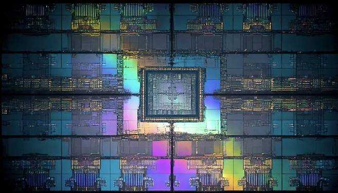 편광 현미경으로 포착한 반도체 다이 단면이다. 중앙의 암청색 코어를 중심으로 무지개빛 간섭색이 방사형으로 퍼져 나가는 구조는 금속 배선층과 절연층이 빛을 굴절·반사하며 만들어낸 물리적 현상이다. 아름답지만 본질은 미학이 아니다. 격자 블록 하나하나가 파라미터가 통과해야 할 감옥 창살이고, 중앙으로 수렴하는 배선망 전체가 곧 지연(latency)의 지도다. 이미지가 아름다울수록 칩 안에서 데이터가 넘어야 할 벽도 많다는 뜻이다. / 해설 = 김현우 기자