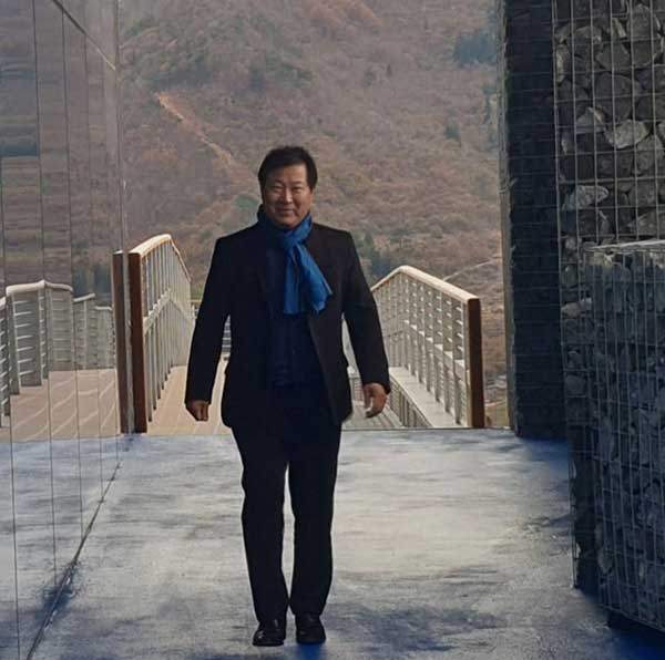 고흥 우주발사전망대 앞바다에 옥빛과 딥블루가 교차하는 비현실적인 바다 위의 섬 옥대도에 도착한 뒤 해안가로 걸어가는 진 회장. 그는