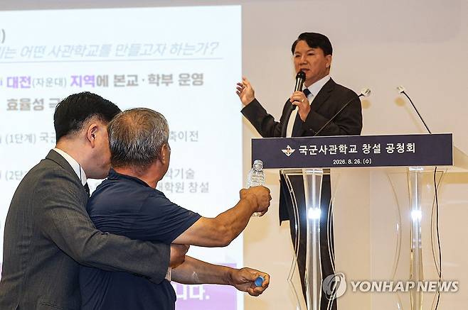 국군사관학교 창설 공청회 (서울=연합뉴스) 최재구 기자 = 26일 서울 용산구 국방컨벤션에서 열린 국군사관학교 창설 공청회에서 한 참석자가 발언하고 있는 김홍철 국방부 정책실장에게 생수병을 들고 다가서다 저지당하고 있다.  2026.8.26  [공동취재] jjaeck9@yna.co.kr
