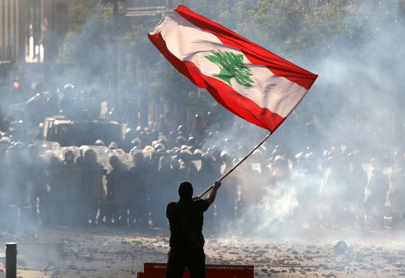 레바논 수도 베이루트에서 8일(현지시간) 벌어진 반정부 시위에서 최루탄 연기가 자욱한 가운데 한 시민이 백향목 문양이 그려진 대형 레바논 국기를 흔들며 시위 진압 경찰과 대치하고 있다.베이루트 로이터 연합뉴스