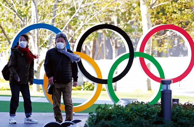 3월16일 일본 도쿄의 올림픽 오륜 조형물 앞을 마스크를 쓴 시민들이 지나가고 있다. ⓒEPA 연합