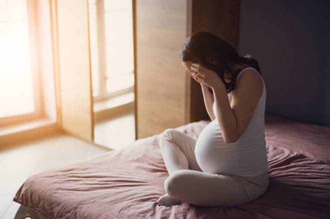 Pregnant woman is having headache. She is sitting on bed at home.
