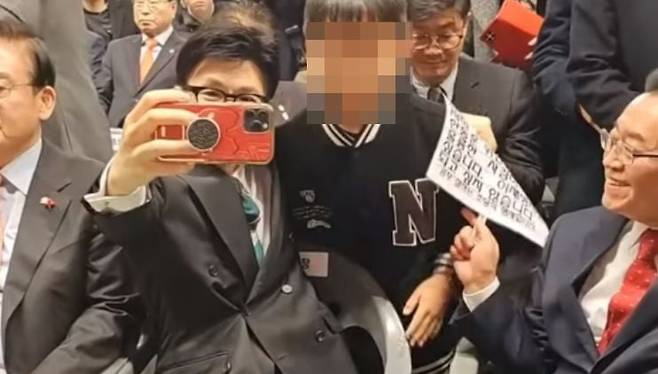 한동훈 국민의힘 비상대책위원장이 지난 4일 충북도당 신년 인사회에서 한 어린이와 사진을 촬영하고 있다. 아이가 준비해온 피켓 속 문구가 아이가 들고 있기에는 부적절하다고 느낀 한 위원장은 이종배 의원이 재차 피켓을 사이로 들이밀자 아이 어깨를 감싼 왼손으로 피켓을 뺏어 등 뒤로 숨기고 있다. / 사진=유튜브 '김사랑 시인' 캡처