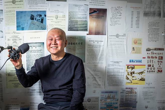 김성민 자유북한방송 대표가 지난 8일 강화도 집에서 방송용 마이크를 잡고 있다. 벽면에는 유튜브 방송 등에 활용할 생각으로 북한 사람들한테 받은 노동당 내부 자료를 빼곡히 붙여 놓았다. 항암 주사를 맞는 탓에 머리는 모두 빠졌고, 정수리 가까이엔 2017년 뇌종양 수술의 흉터가 10cm가량 남았다. /이신영 영상미디어 기자