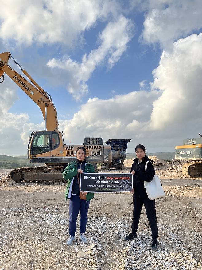 팔레스타인평화연대의 자아 활동가(오른쪽)가 뎡야핑 활동가와 함께 2023년 4월13일 팔레스타인 서안지구 남쪽에 있는 알나캅(네게브) 지역 유대인 정착촌 건설 현장에서 에이치디(HD)현대 굴착기를 발견하고 미리 준비한 펼침막을 그 앞에서 들고 있다. 자아 활동가 제공