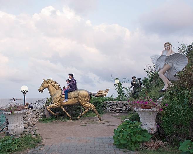 강원도 고성의 한 카페에서 철책선 앞 말 조형물에 올라가 있는 여행객들, 뒤로 군인과 마릴린 먼로 조형물도 함께 있다. 2018년/ 사진가 김전기