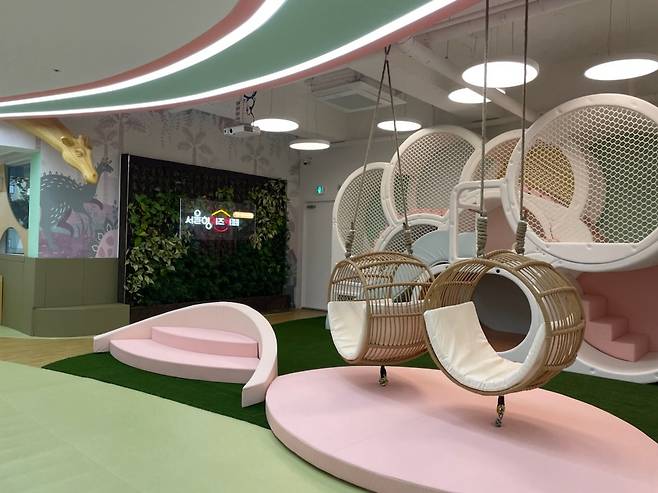 실내 놀이터에 흔들 그네와 미끄럼틀이 설치되어 있는 사진이다. 벽면에는 하하호호 놀이터 로고와 수직 정원이 조성되어 있다.