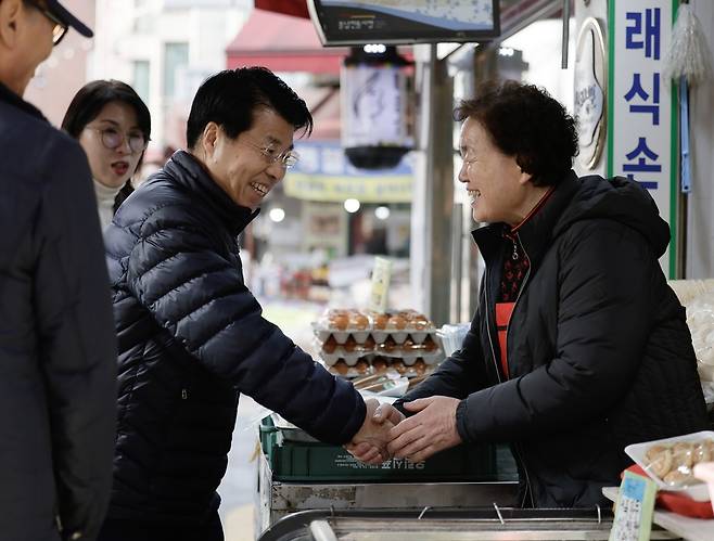 서강석 송파구청장이 시장 상인과 악수하며 미소짓고 있는 사진이다. 상점 앞에는 달걀과 만두가 놓여 있다.